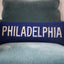 Navy blue lumbar pillow with white embroidered text reading PHILADELPHIA, displayed on a gray upholstered chair