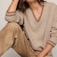 A woman wearing a beige v-neck knit sweater with striped cuffs and matching beige pants, posed against a light neutral studio background.