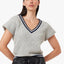 A woman modeling a light gray ribbed knit V-neck sweater with navy blue trim, paired with black pants, against a plain light gray background.