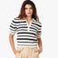 Woman wearing navy and cream striped polo sweater with short sleeves and beige trousers, standing against white background