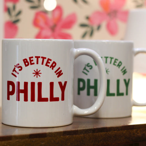Two white ceramic mugs with red and green text reading It's Better In Philly, displayed on wooden surface with pink floral b…