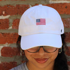 Person wearing a white cap with an American flag design against a brick wall.