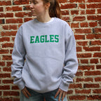 Woman wearing light blue crewneck sweatshirt with green EAGLES text, standing against red brick wall