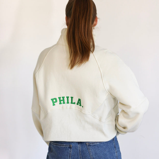 A person shown from the back wearing an off-white cropped sweatshirt with green 'PHILA.' lettering on the back, paired with blue jeans, against a plain white background. Main image