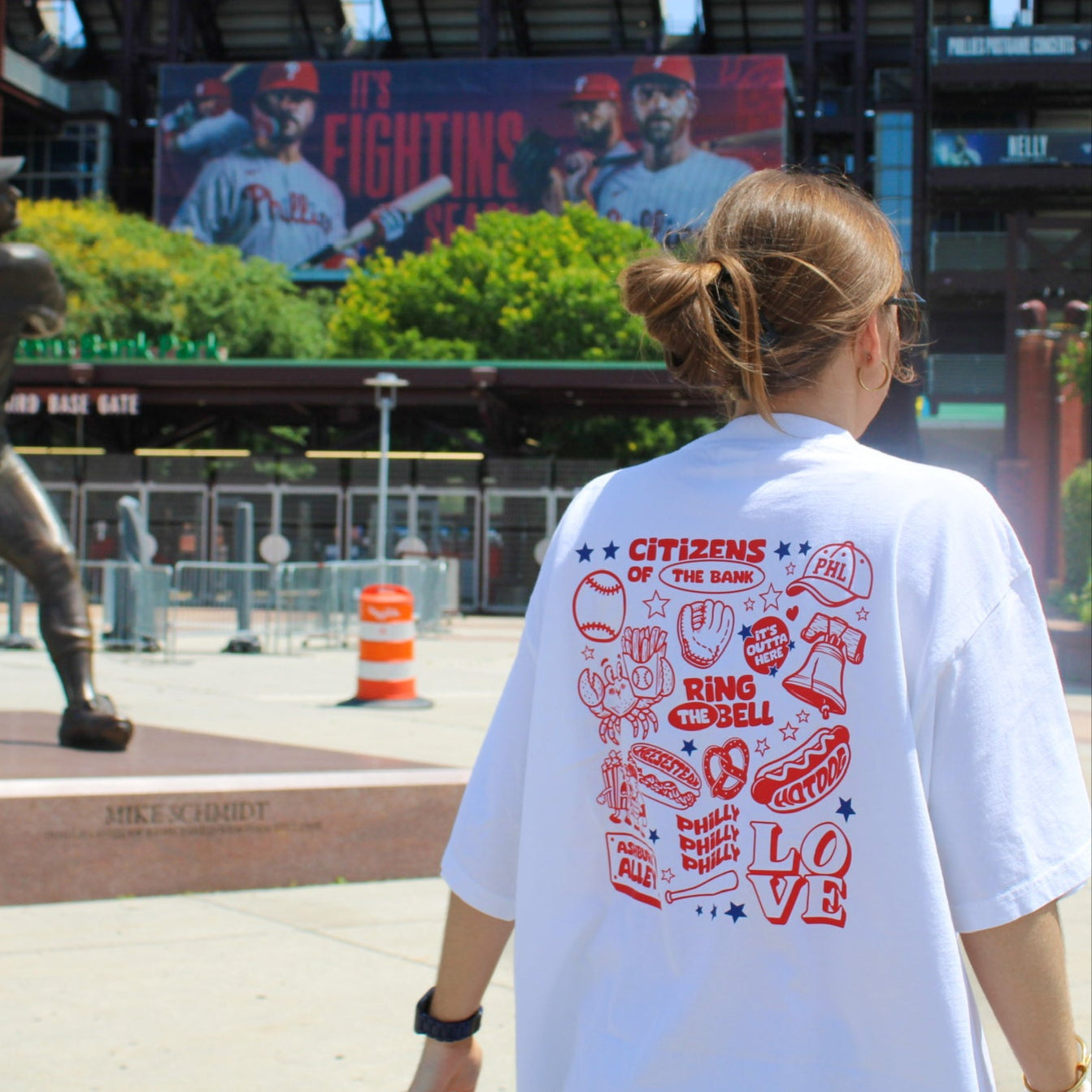 Person wearing a white t-shirt with red graphics and text, standing in front of a sports stadium with a large screen displaying athletes.