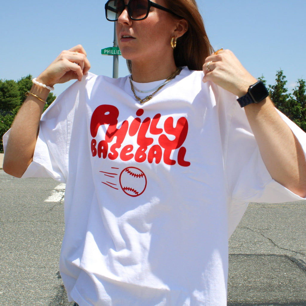 Person wearing a white t-shirt with 'Philly Baseball' text and graphic outdoors.