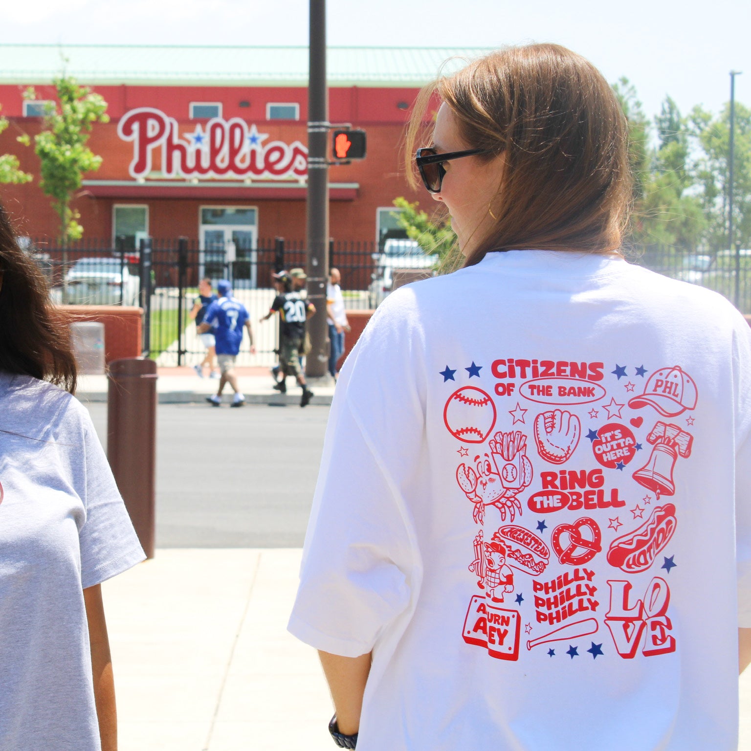 Person wearing a white t-shirt with red graphics and text, standing in front of a Phillies building.