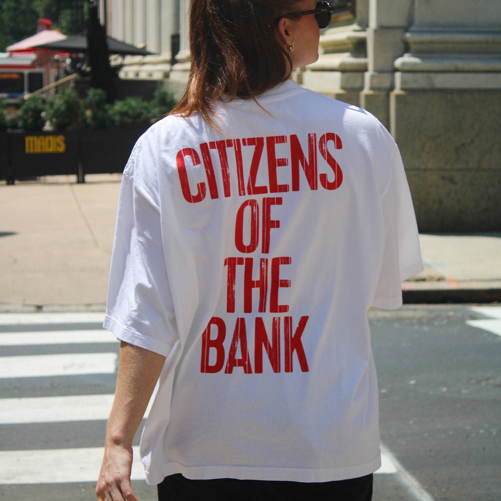 Woman wearing white drop shoulder tee with red text reading Citizens of the Bank, standing outdoors on city street