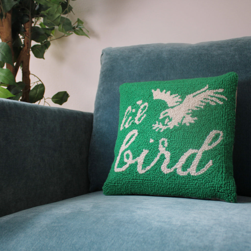 Green hooked pillow with cream bird design and 'Li'l Bird' text on blue upholstered sofa