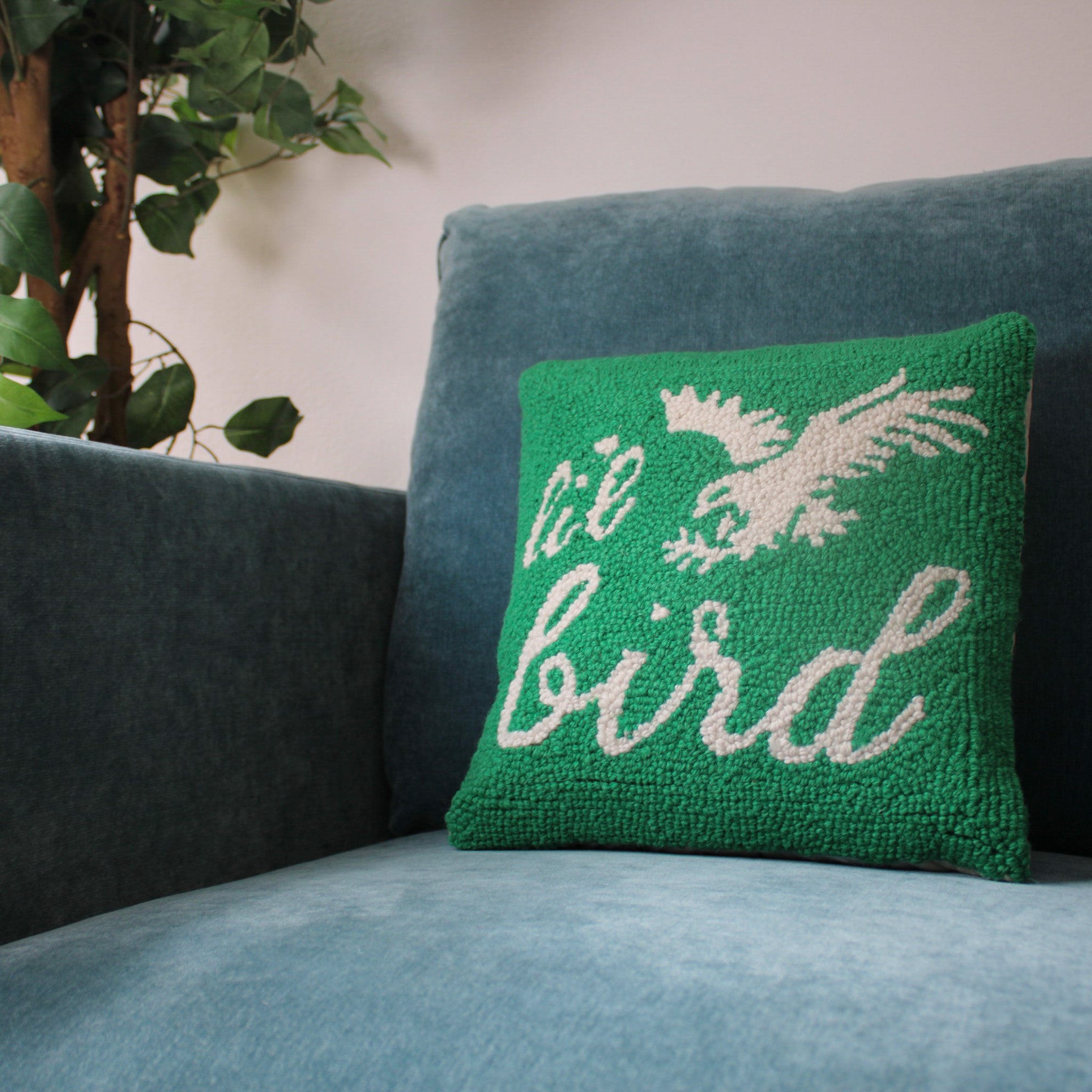 Green hooked pillow with cream bird design and 'Li'l Bird' text on blue upholstered sofa