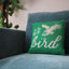 Green hooked pillow with cream bird design and 'Li'l Bird' text on blue upholstered sofa