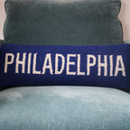 Navy blue lumbar pillow with white embroidered text reading PHILADELPHIA, displayed on a gray upholstered chair