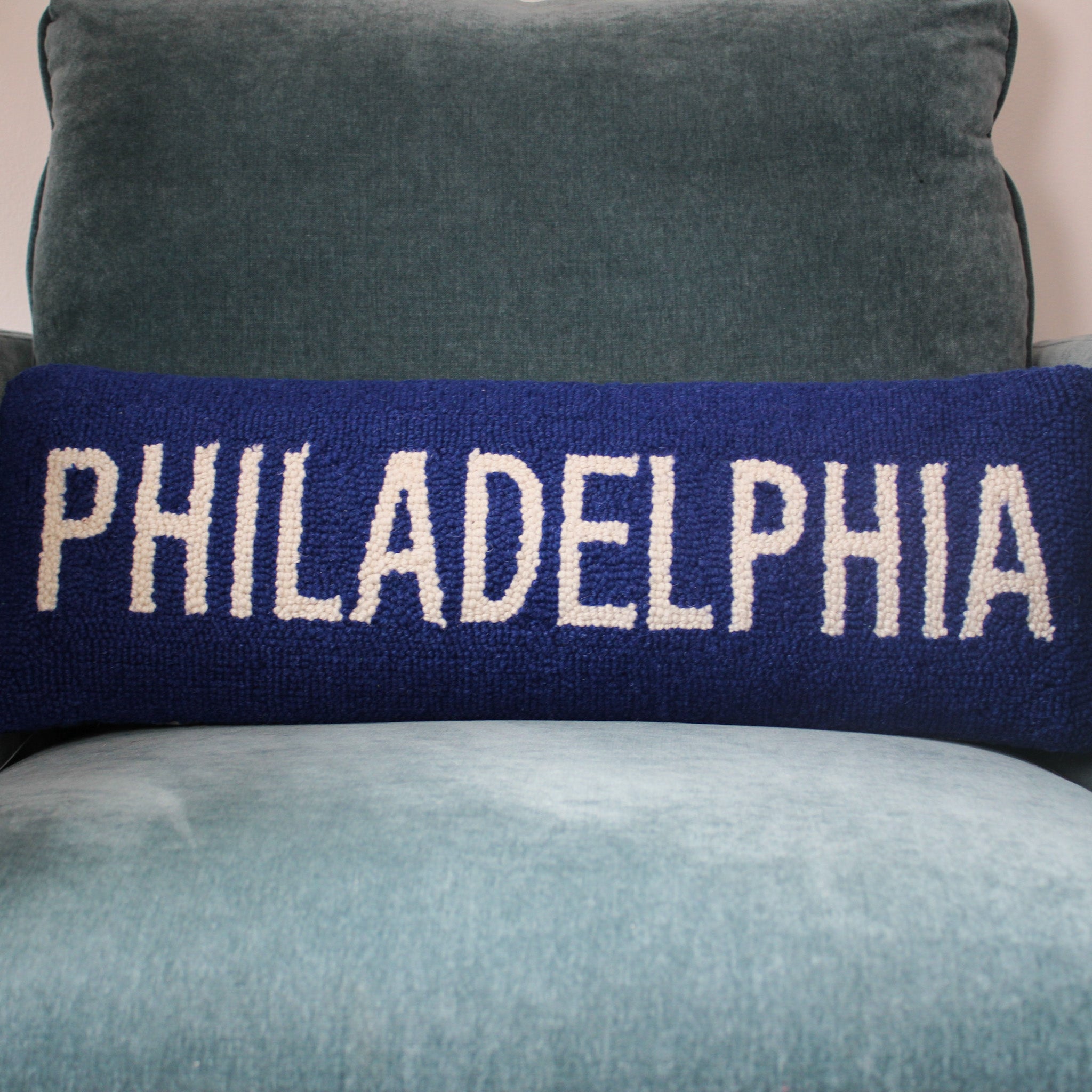 Navy blue lumbar pillow with white embroidered text reading PHILADELPHIA, displayed on a gray upholstered chair