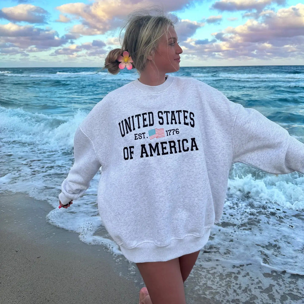 Woman wearing light gray United States of America embroidered crew sweatshirt at the beach with ocean waves in background