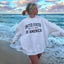 Woman wearing light gray United States of America embroidered crew sweatshirt at the beach with ocean waves in background