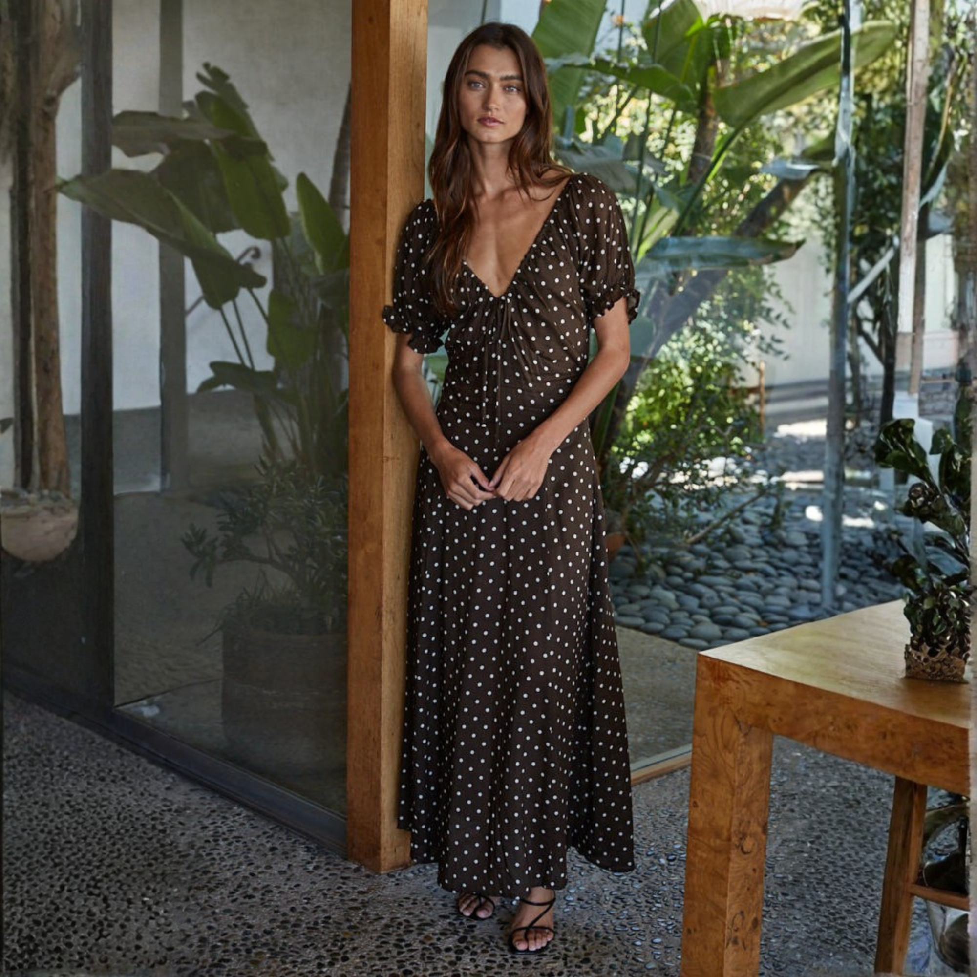 Woman wearing black polka dot sweetheart neckline midi dress with puffed sleeves, standing in modern sunlit space with plants Main image