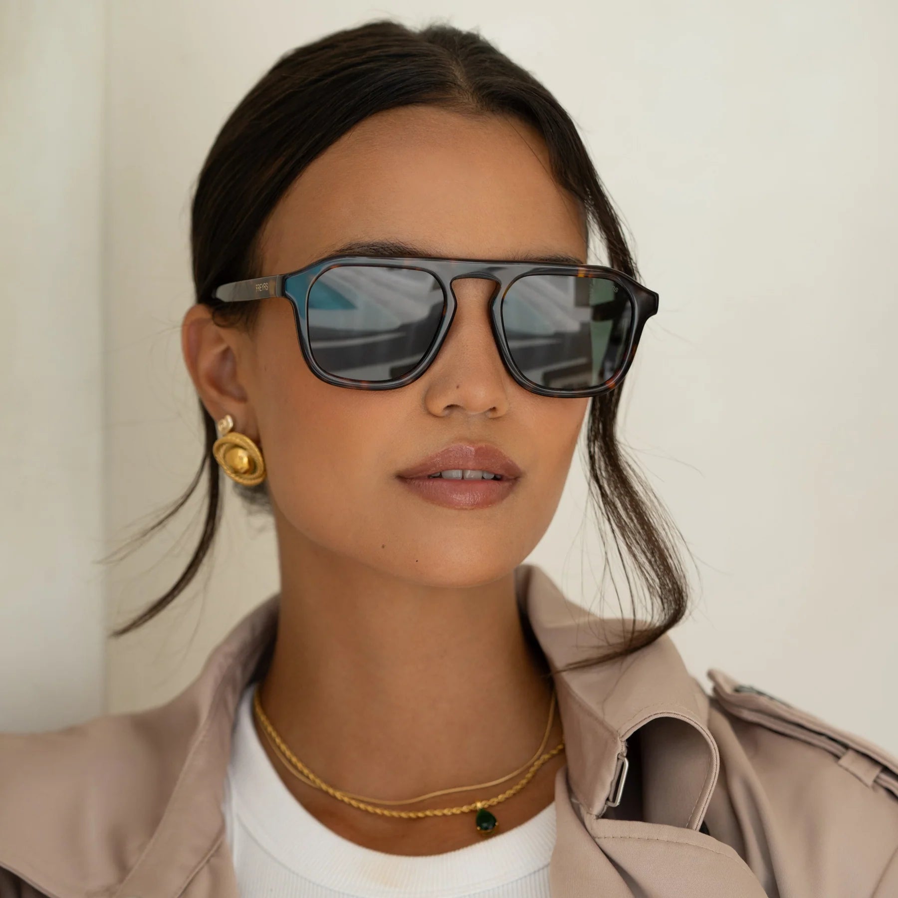 Woman wearing Addison sunglasses with dark tinted lenses and tortoiseshell frame, paired with gold hoop earrings and beige b…