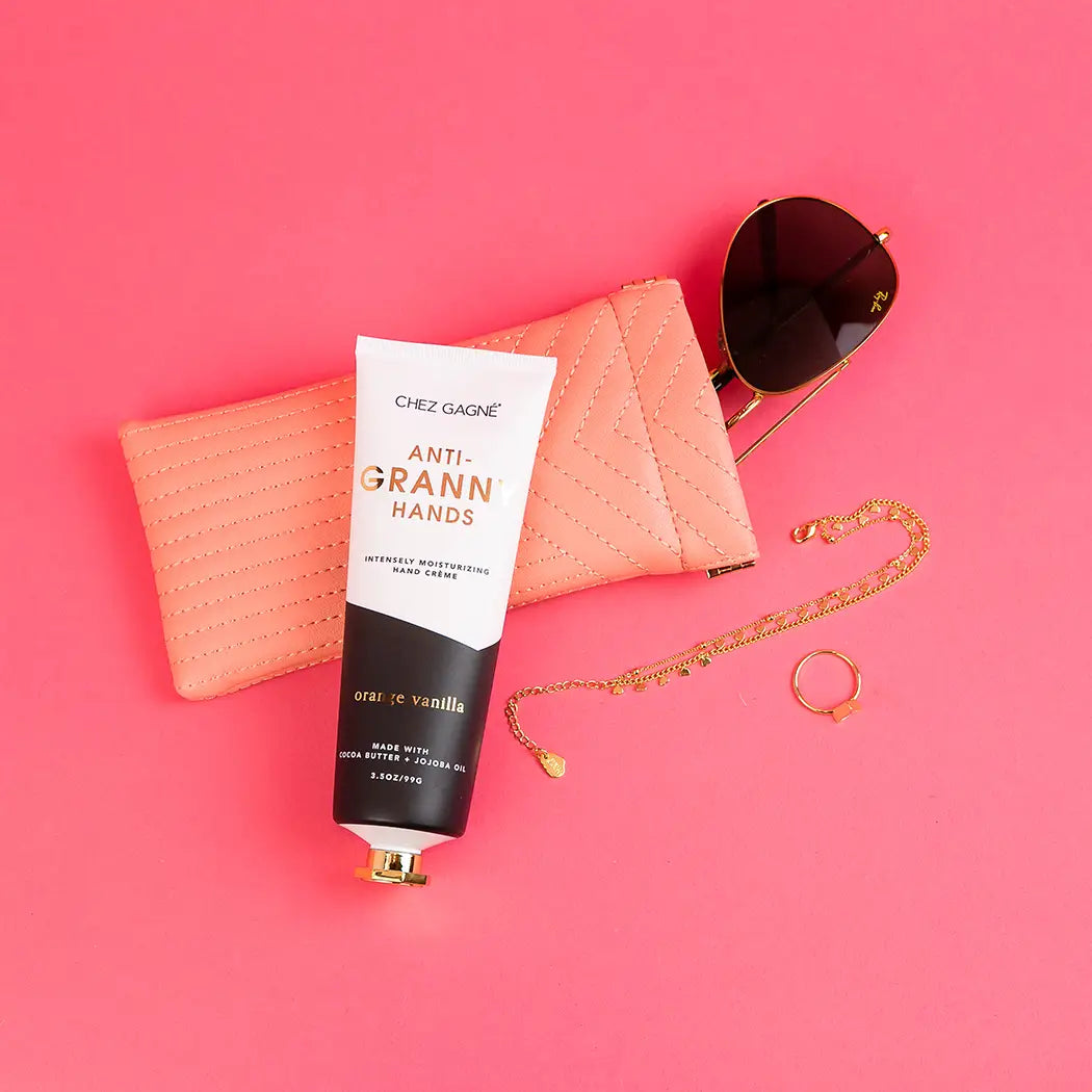Anti-Granny Hands Orange Vanilla hand cream tube displayed with peach quilted wallet, sunglasses, and gold jewelry on pink b… Main image
