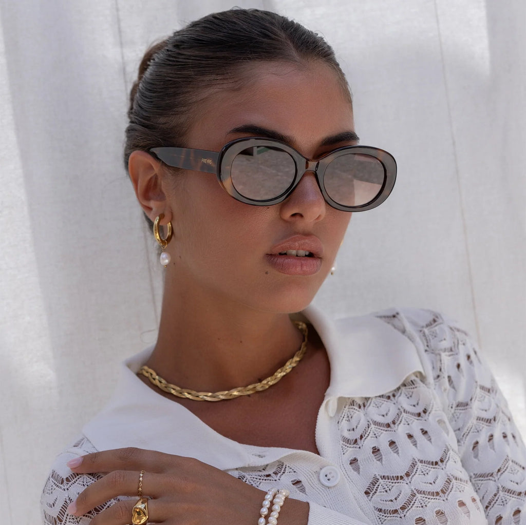Woman wearing oversized round tortoiseshell sunglasses with tinted lenses, gold chain necklace, and white lace blouse