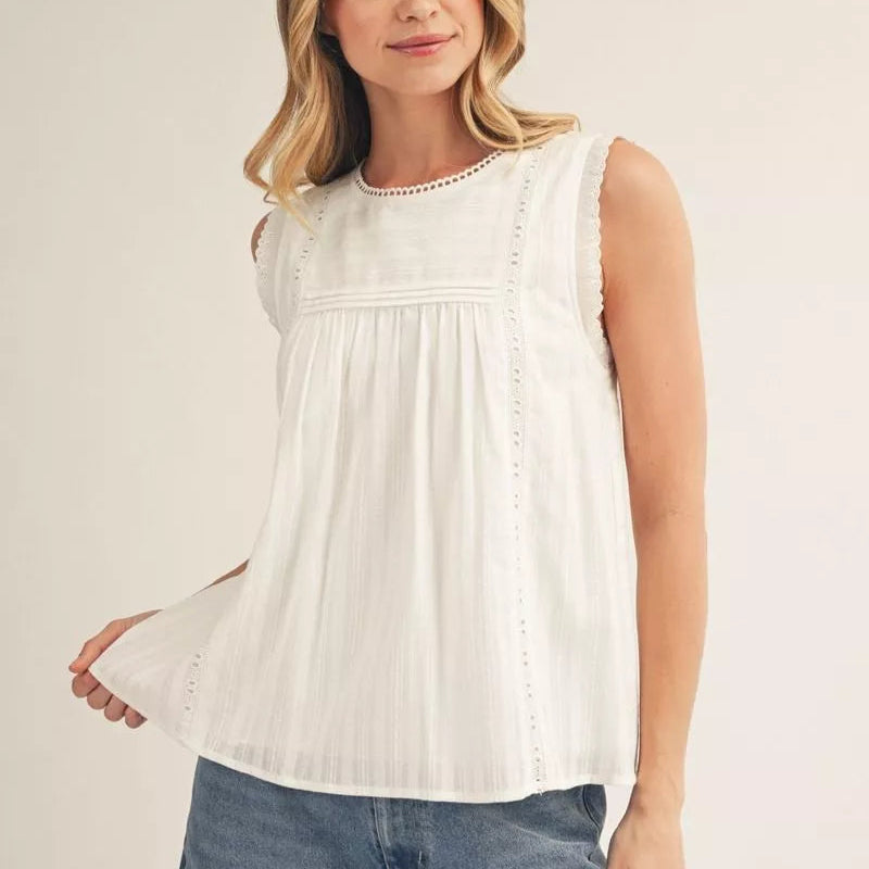 Woman wearing a cream sleeveless babydoll tank top with pleated front and eyelet lace trim, paired with blue jeans