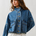 Woman wearing medium-wash denim jacket with balloon sleeves, front button closure, chest pockets, and frayed hem