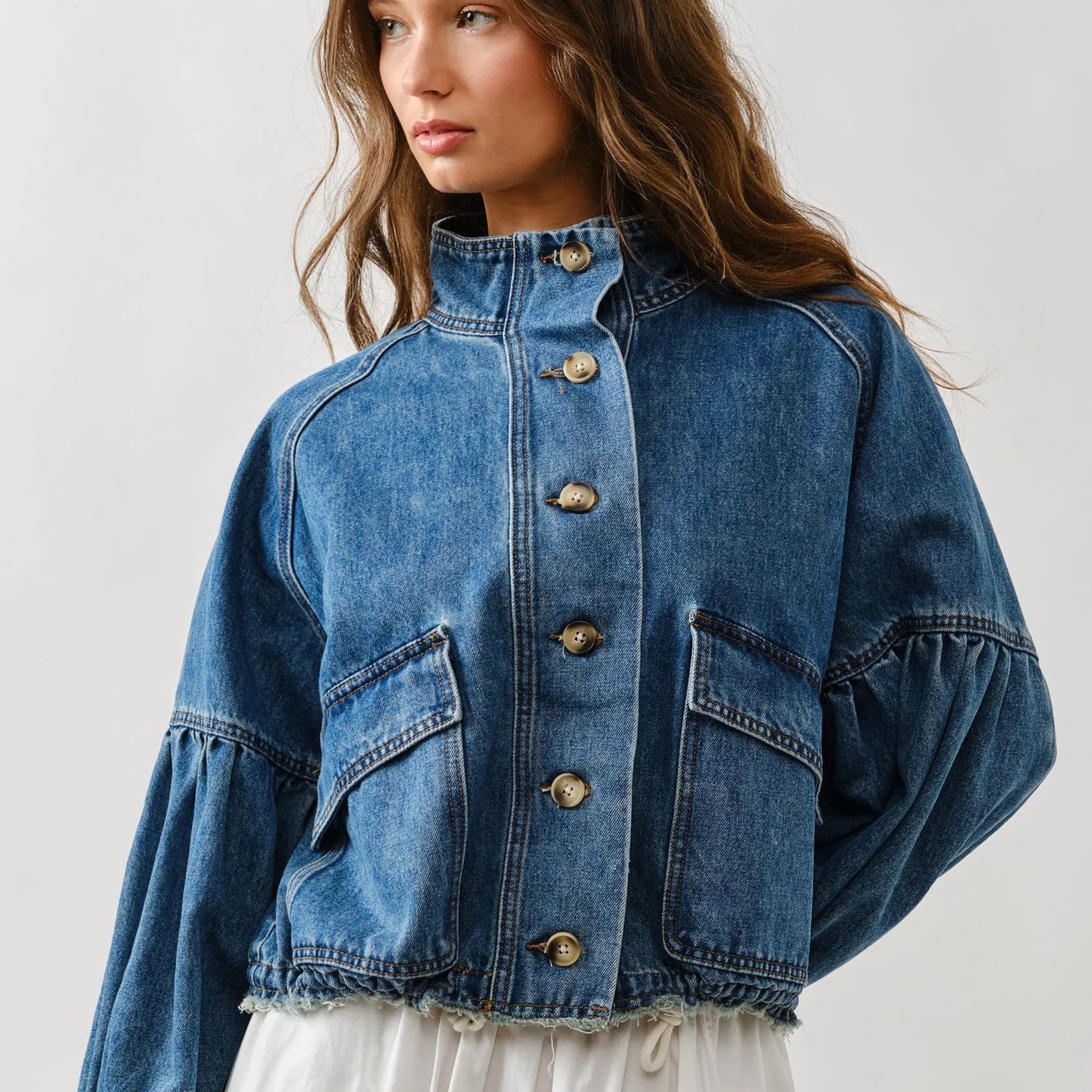 Woman wearing medium-wash denim jacket with balloon sleeves, front button closure, chest pockets, and frayed hem