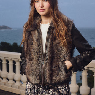 A woman stands on a balcony overlooking the sea, wearing a brown and black faux-fur and leather jacket over a white top and a patterned skirt.
