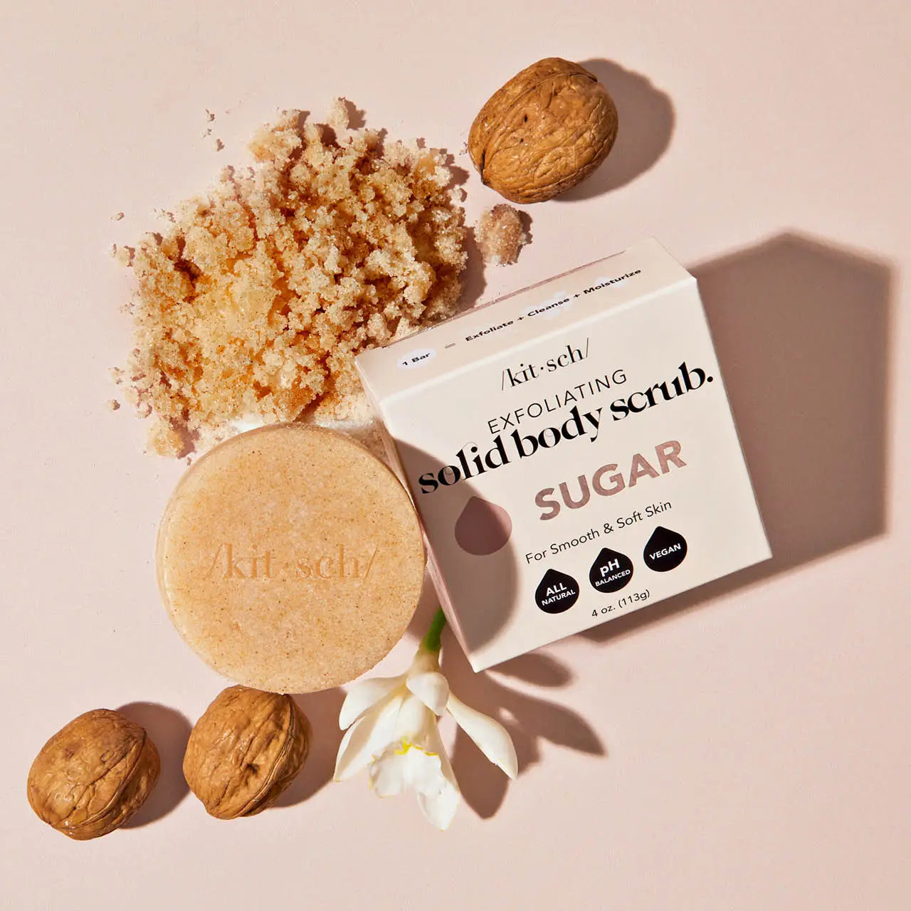 Sugar exfoliating body scrub bar with scattered sugar granules, whole almonds, and white flower petals on cream background