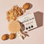 Sugar exfoliating body scrub bar with scattered sugar granules, whole almonds, and white flower petals on cream background