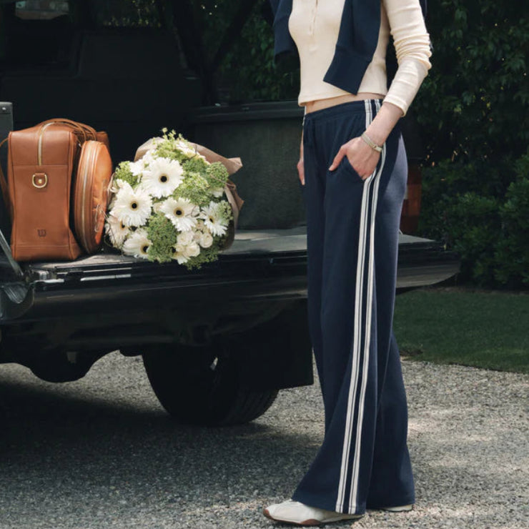 A woman stands outdoors beside the open trunk of a dark vehicle, wearing a navy sleeveless vest and matching wide-leg pants with white side stripes, a cream long-sleeve top, white sneakers, and a navy cap. A brown suitcase and a bouquet of white flowers sit in the truck bed. Main image