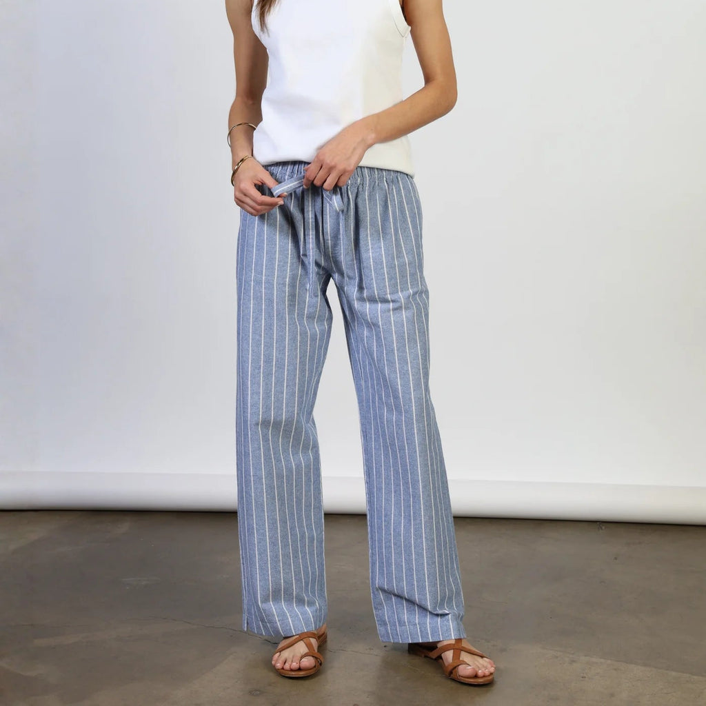 Woman wearing white sleeveless tank top and blue pinstriped linen pants with brown sandals, standing against white wall