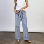 Woman wearing white sleeveless tank top and blue pinstriped linen pants with brown sandals, standing against white wall