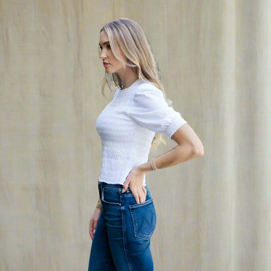 Woman wearing a white top and blue jeans standing outdoors with greenery in the background