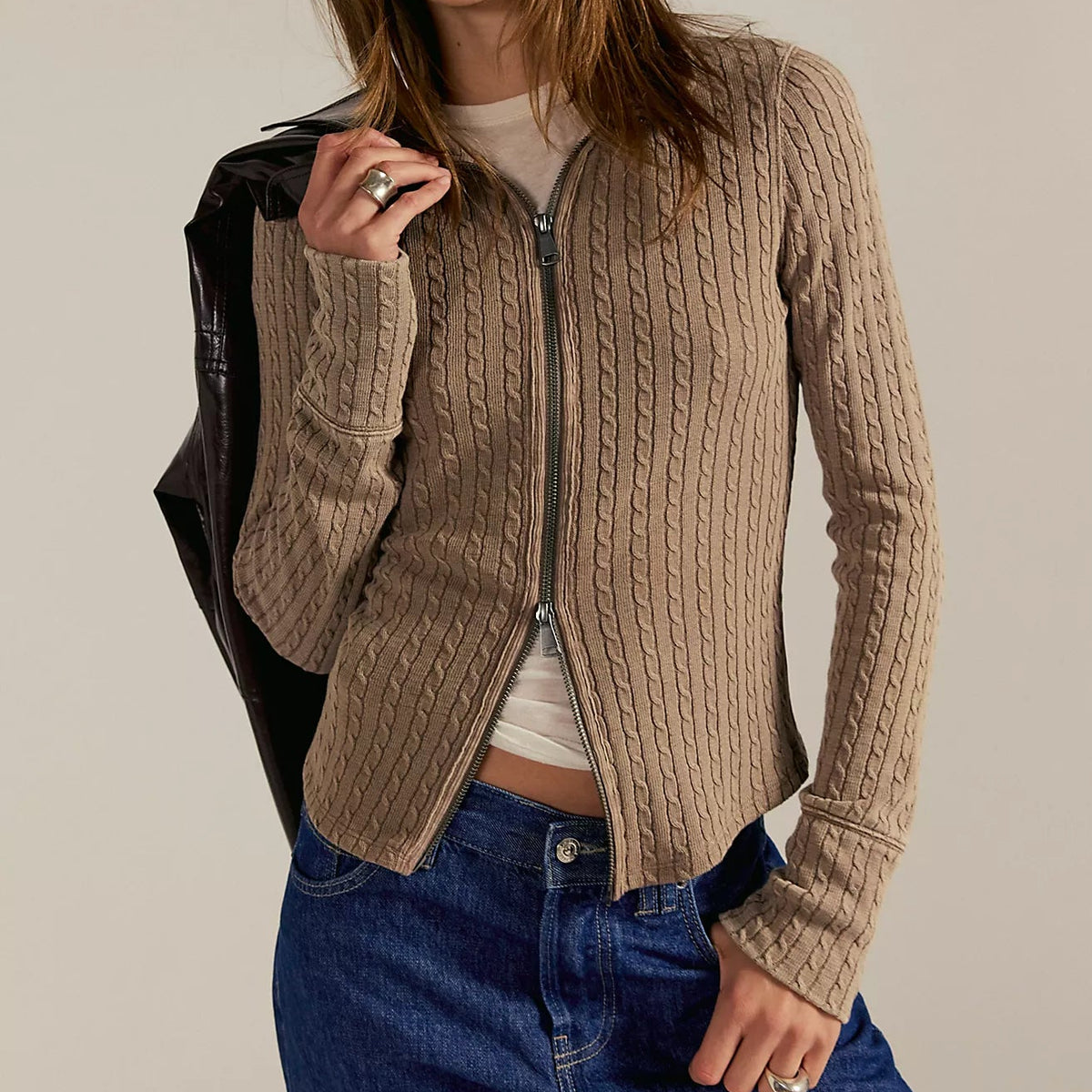 Woman wearing beige cable-knit zip-up cardigan over white tee with dark jeans, holding black leather jacket