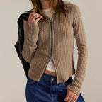 Woman wearing beige cable-knit zip-up cardigan over white tee with dark jeans, holding black leather jacket