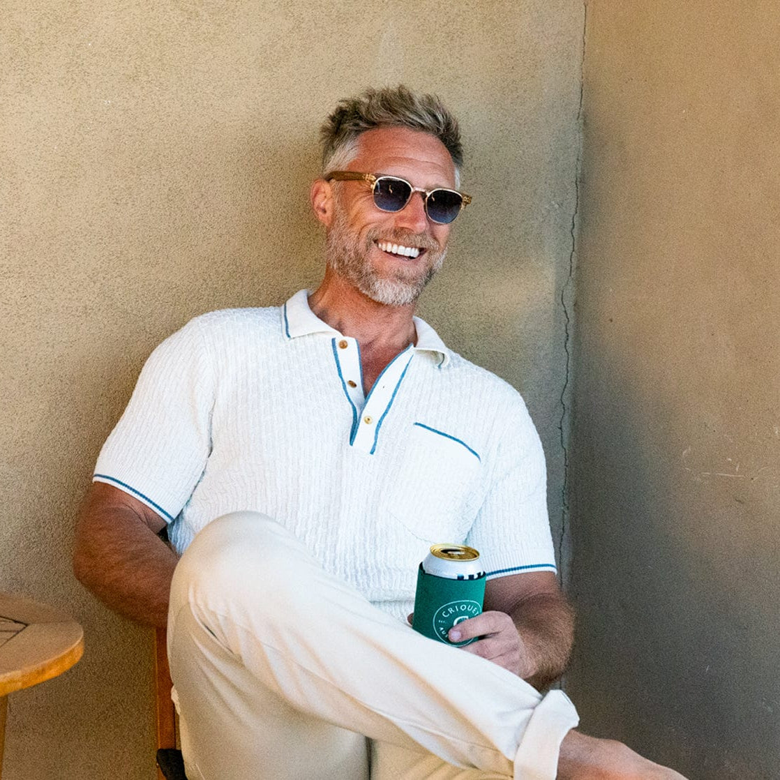 Man sitting against a beige wall holding a can, wearing sunglasses and a white shirt.