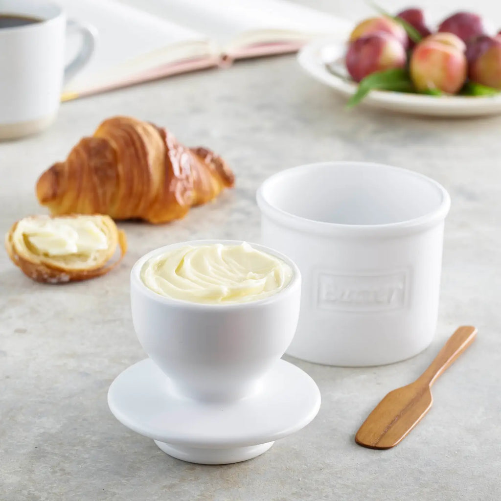 The Original Butter Bell Crock with cream-colored lid containing whipped butter, displayed with croissants and fresh apples …