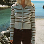 A blonde woman stands on a rocky beach wearing a cropped, long-sleeve knit cardigan with horizontal stripes in light gray, beige, and brown, paired with dark brown pants. A bridge and blue ocean are visible in the background.