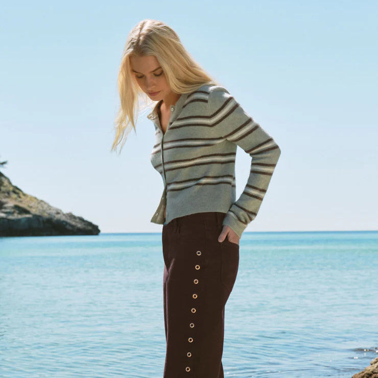 A woman stands barefoot on a rocky shoreline with the ocean and sky behind her, wearing a gray and beige striped long-sleeve top and dark brown wide-leg pants with a vertical row of silver-tone eyelet buttons down the side.