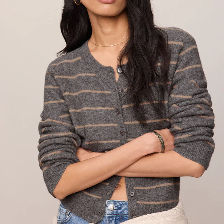 A woman modeling a gray and beige striped knit cardigan with a button-up front, paired with blue jeans and a cream sweater tied around her waist, against a solid light beige background.