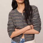 A woman modeling a gray and beige striped knit cardigan with a button-up front, paired with blue jeans and a cream sweater tied around her waist, against a solid light beige background.