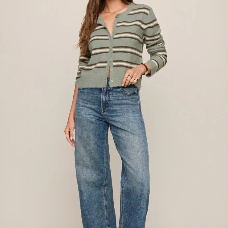 A woman modeling a light sage green and beige striped knit cardigan with a split neckline, paired with wide-leg blue jeans and brown boots, against a solid light beige studio background.