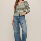 A woman modeling a light sage green and beige striped knit cardigan with a split neckline, paired with wide-leg blue jeans and brown boots, against a solid light beige studio background.