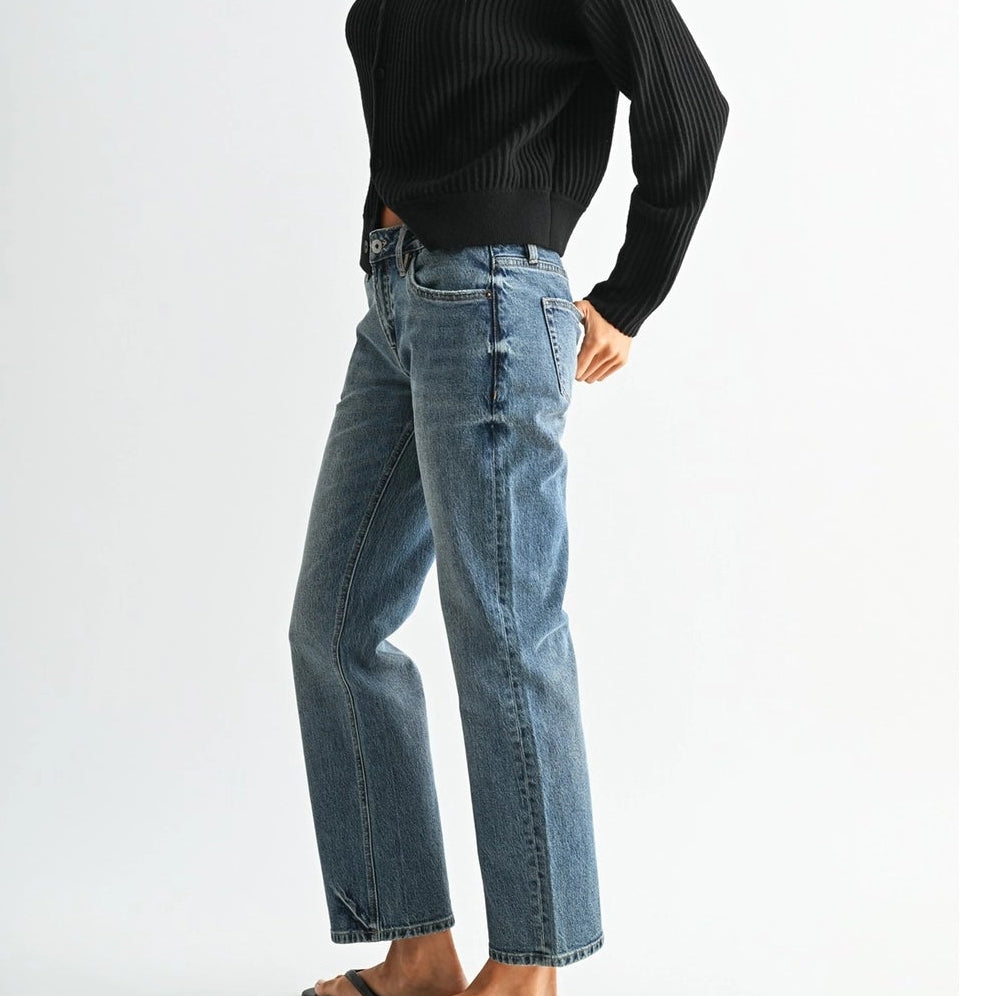 A woman wearing a black ribbed long-sleeve turtleneck sweater, light-wash blue jeans, and black flip-flops, standing in profile against a plain light gray studio background.