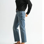 A woman wearing a black ribbed long-sleeve turtleneck sweater, light-wash blue jeans, and black flip-flops, standing in profile against a plain light gray studio background.