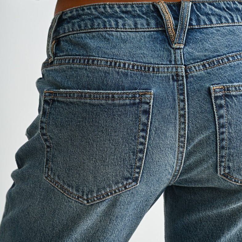 Close-up back view of a person wearing blue denim jeans with a black ribbed top, showing the waistband, belt loops, and back pockets against a light gray studio background.
