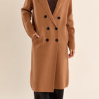 A woman modeling a camel brown double-breasted wool coat over a dark brown V-neck sweater, set against a solid light beige background.