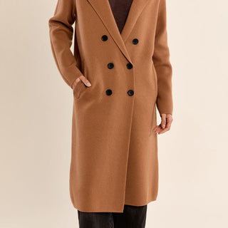 A woman modeling a camel brown double-breasted wool coat over a dark brown V-neck sweater, set against a solid light beige background.