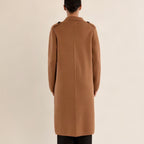 A model viewed from the back wearing a long camel brown coat with shoulder epaulets and black trousers, set against a solid light beige background.