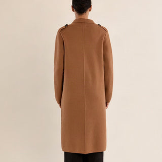 A model viewed from the back wearing a long camel brown coat with shoulder epaulets and black trousers, set against a solid light beige background.